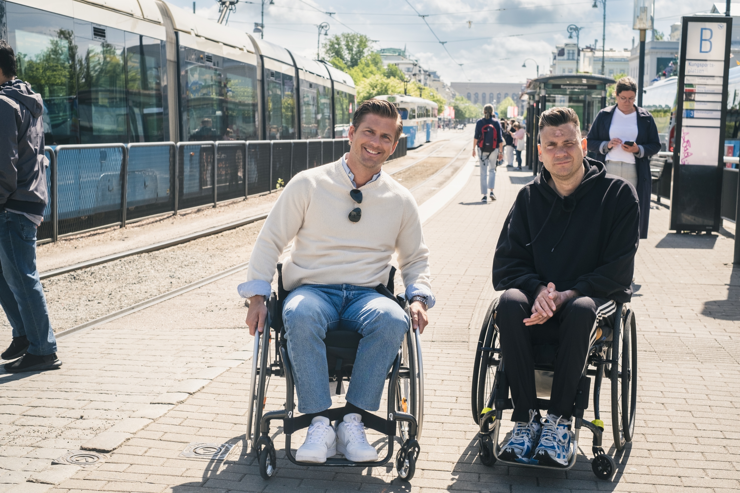 Hampus Magnusson och Anders Westgerd, båda sitter i rullstol på hållplats med spårvagnar i bakgrunden.